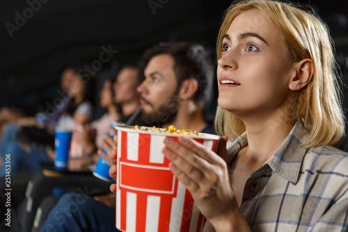 Side view of shocked beautiful blonde woman watching excited horror in cinema hall. Girl in checkered shirt keeping popcorn and looking at screen. Concept of movie time.