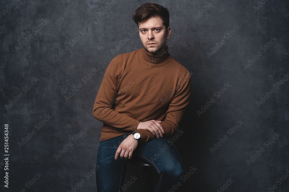 handsome casual man sits on chair near a dark gray wall and looks to side, portrait picture.