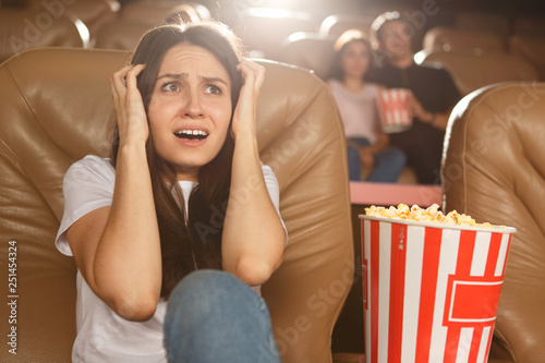Frightened girl watching terrible horror movie in cinema. Dark haired young woman in white shirt and jeans sitting on comfortable chair and closing ears with hands. Concept of horror films.