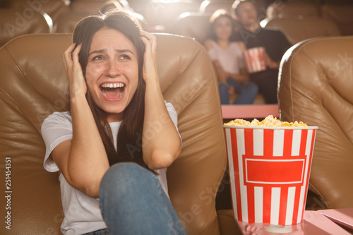 Pretty brunette woman in white shirt and jeans sitting on comfortable chair and closing ears with hands. Frightened girl watching terrible horror movie in cinema. Horror films concept.