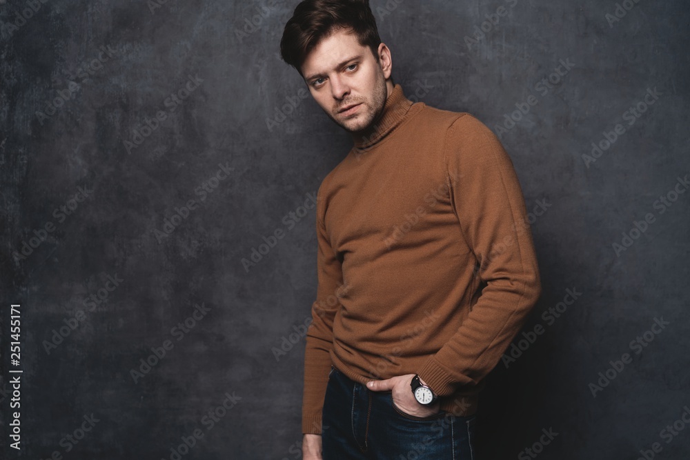 Handsome young man on gray background looking at camera. Portrait of young man with hands in pockets leaning against gray wall.