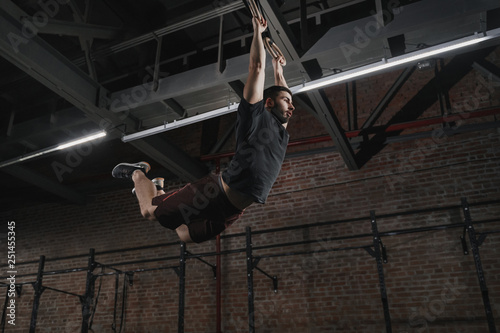 Young crossfit athlete swinging on gymnastic rings doing pull-ups at the gym. Workout exercises © Arsenii