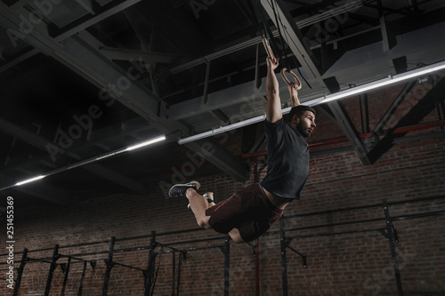 Young athlete doing pull-ups on gymnastic rings at crossfit gym. Handsome man practicing muscle ups swinging workout exercise.