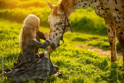 mother with a little girl in dresses stroke a spotted horse