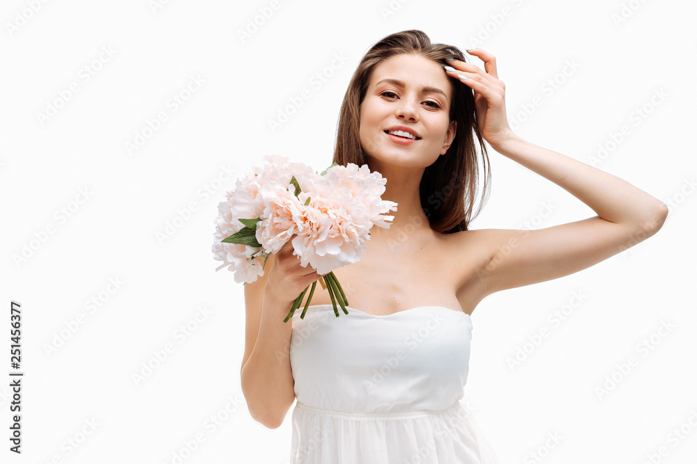 Portrait beautiful young girl smiling and posing with artificial flowers on white background in white dress.