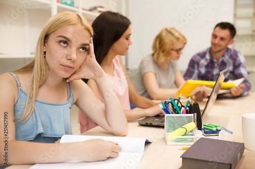 Blonde student girl sitting at table and writing lecture in notebook   looking up and wanting sleep. Group of students sitting at table  studying and doing homework using notes and laptop in classroom