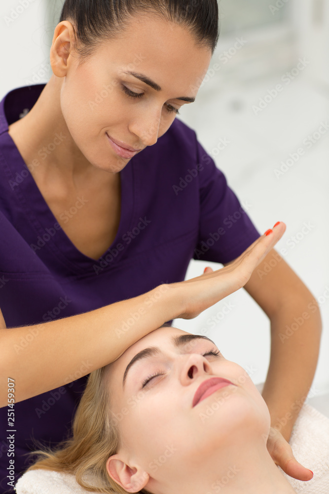 Young smiling cosmetologist stroking face of female client with massage movements. Beautiful blonde lying on couch and relaxing during rejuvenating procedure. Concept skin care and beauty. Young smiling cosmetologist stroking face of female client with massage movements. Beautiful blonde lying on couch and relaxing during rejuvenating procedure. Concept skin care and beauty.