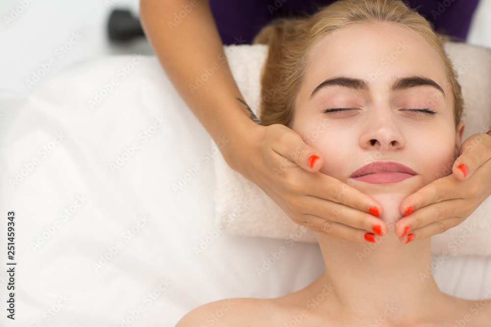Close up of woman face lying on couch and enjoying relaxing lifting procedure in spa salon. Hands of masseuse massaging skin of attractive blonde with closed eyes. Concept of beauty.
