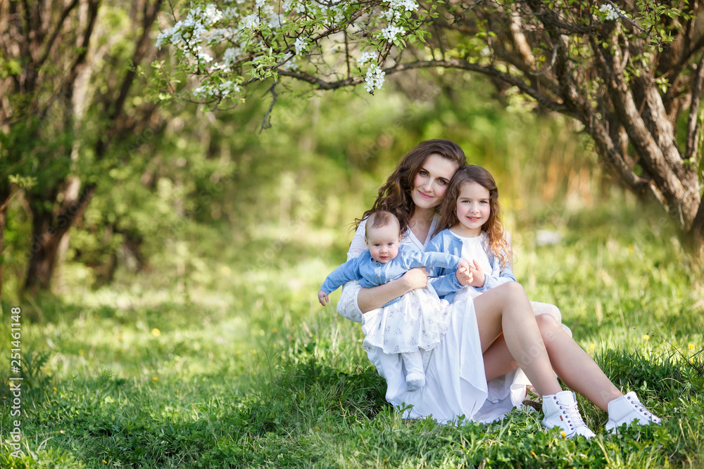 Portrait of young beautiful mother with two daughters near blossoming tree. Mother, two daughters in a spring garden. Portrait of young beautiful mother with two daughters near blossoming tree. Mother, two daughters in a spring garden.