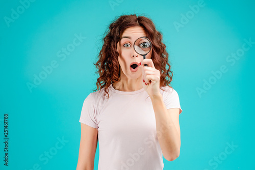 Attractive brunette girl with curly hair isolated over blue turquoise background looking at something through magnifying glass with a big eye. Vision concept