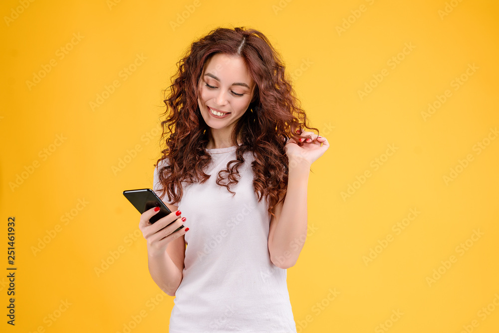 Fototapeta premium Attractive brunette girl with curly hair isolated over yellow background using mobile app for communication