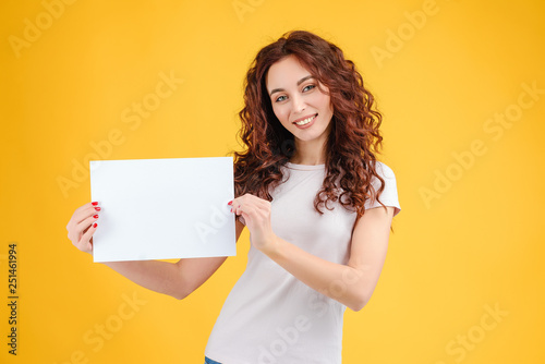 Attractive brunette girl with curly hair isolated over yellow background with a copyspace white paper in her hands