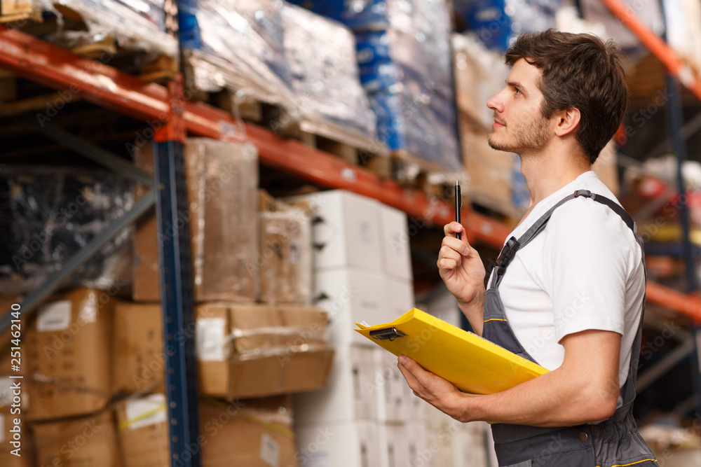 Side view of handsome worker concentrated on goods, standing near shelves in warehouse. Man holding pen and yellow clipboard. Specialist wearing white t shirt and uniform.