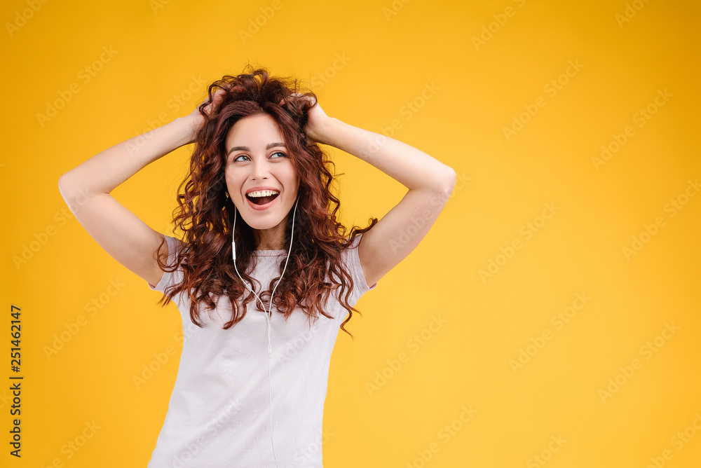 Attractive brunette girl with curly hair isolated over yellow background having fun, listening to music, singing and dancing with earpods on. Music app and online karaoke concept - obrazy, fototapety, plakaty 