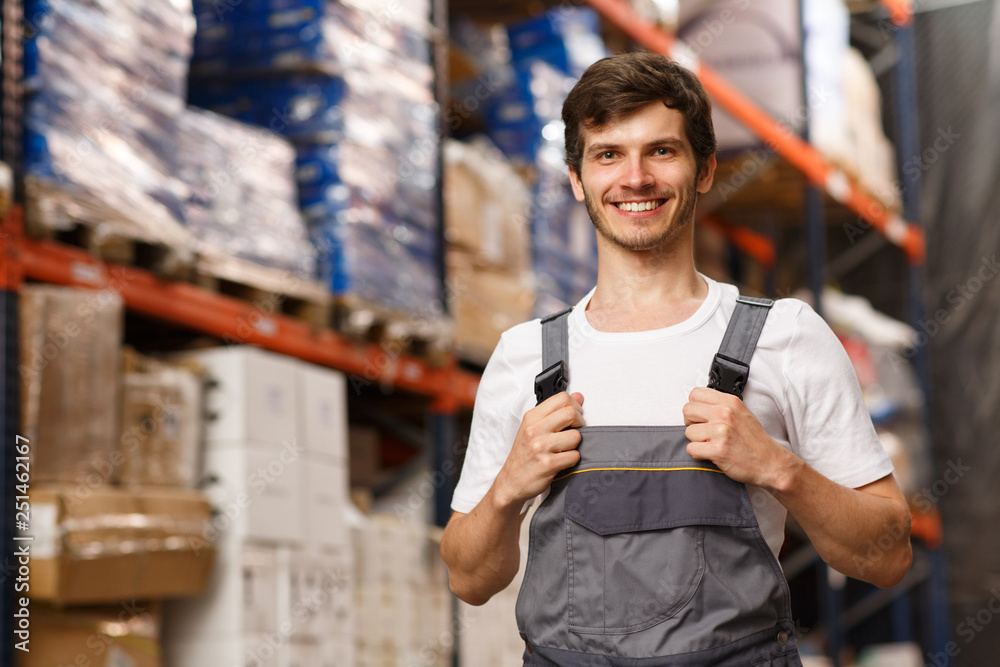 Good looking loader of warehouse posing, smiling and looking at camera. Cheerful worker standing and holding uniform by hands. Forklift and shelves with goods on background. - obrazy, fototapety, plakaty 