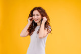 Attractive brunette girl with curly hair isolated over yellow background having fun, listening to music, singing and dancing with earpods on. Music app and online karaoke concept