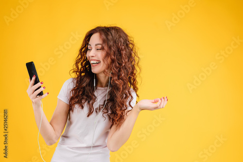 Attractive brunette girl with curly hair isolated over yellow background having fun, listening to music, singing and dancing with cellphone and earpods on. Music app and online karaoke concept