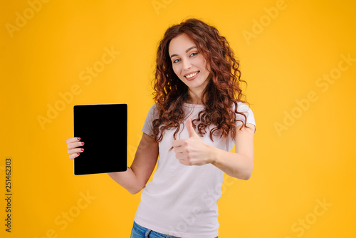Attractive brunette girl with curly hair isolated over yellow background pointing on a copyspace on the tablet in her hand. 