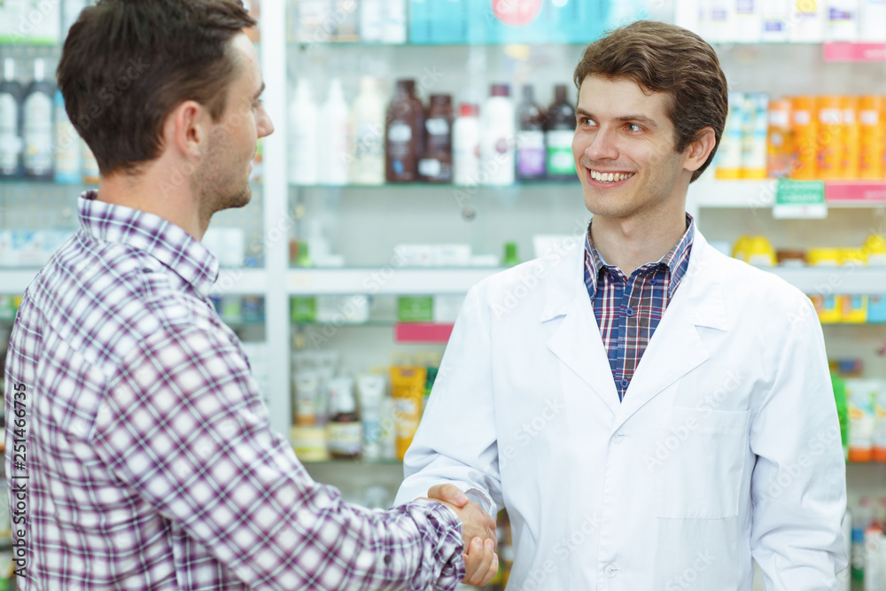 Male pharmacist standing, looking at customer and smiling. Man in checked shirt shaking hand with worker of drugstore. Concept of pharmaceutical industry and medications.