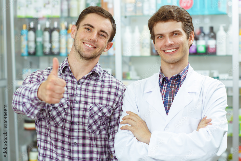 Two good looking men standing in drugstore smiling and posing. Handsome pharmacist in white coat looking at camera and male customer in checked shirt showing thumb up. Two good looking men standing in drugstore smiling and posing. Handsome pharmacist in white coat looking at camera and male customer in checked shirt showing thumb up.