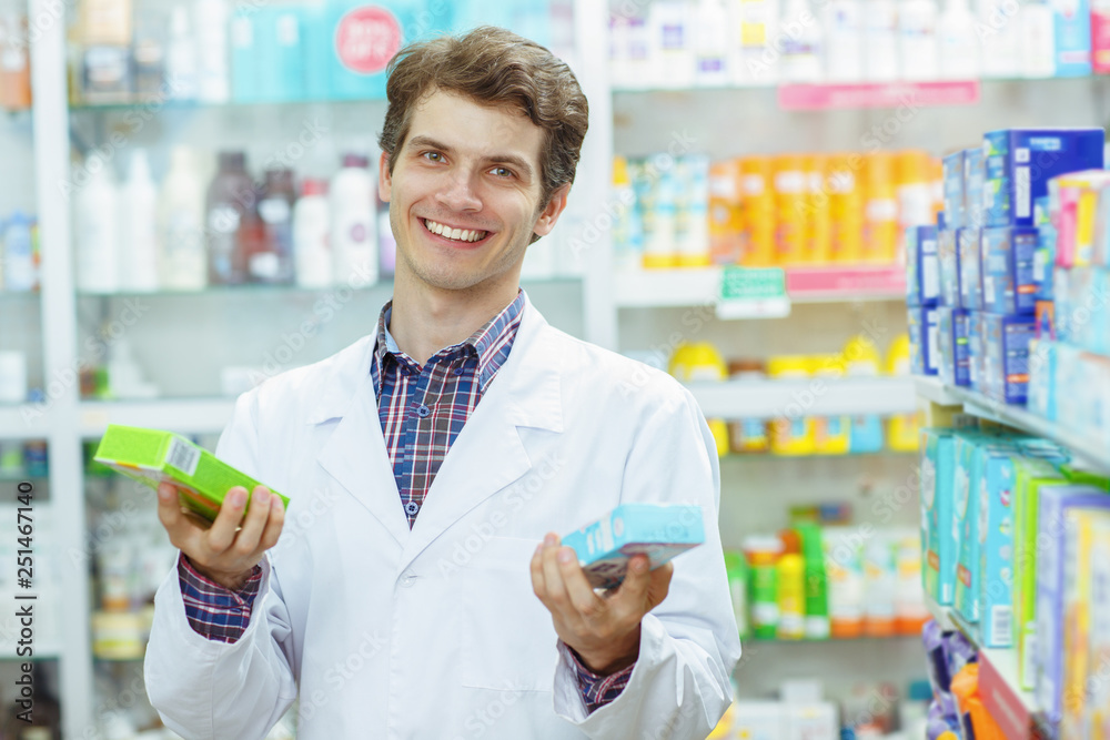 Cheerful male pharmacist standing and holding in hands two different medicaments. Good looking medical consultant in white coat smiling, looking at camera and posing.