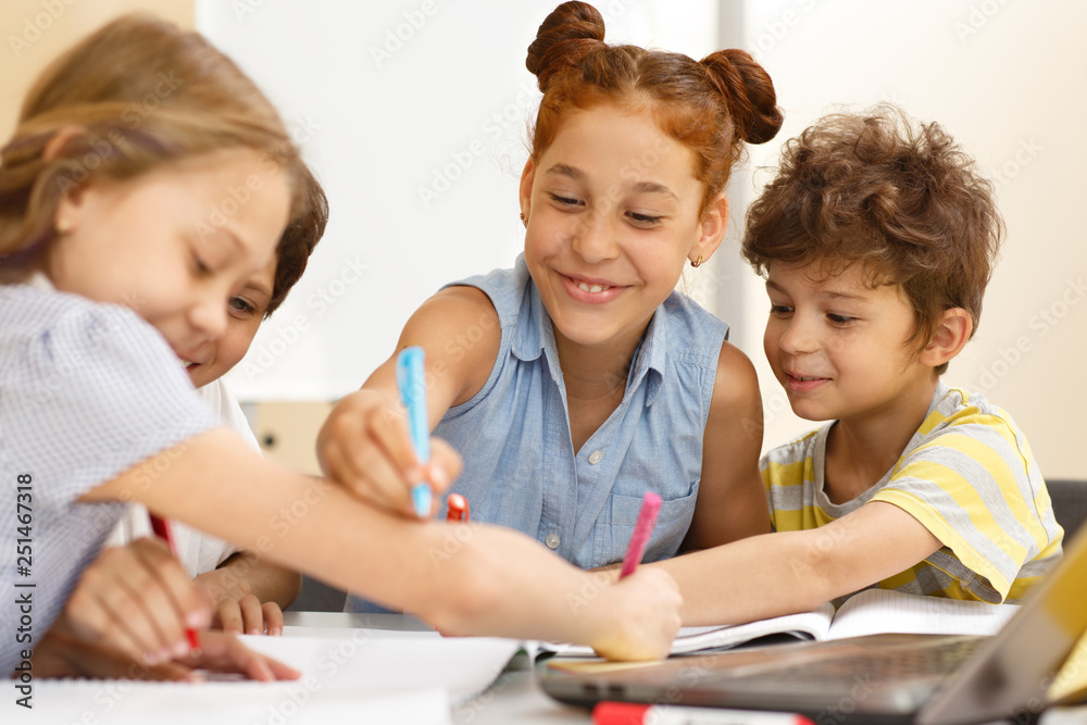 Cheerful schoolgirl sitting at table near friends and drawing with pen on hand of classmate. Happy pupils laughing while working in groups. Concept of interesting lessons and friendly atmosphere. Cheerful schoolgirl sitting at table near friends and drawing with pen on hand of classmate. Happy pupils laughing while working in groups. Concept of interesting lessons and friendly atmosphere.