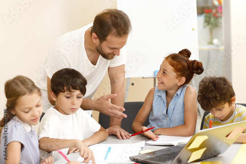 View of happy male teacher in white shirt helping and checking works of pupils in classroom. Diligent little students of school writing, carefully doing tasks and talking with teacher.