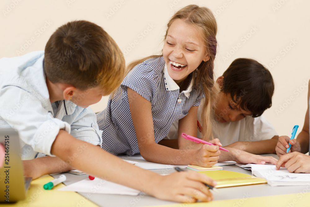 Cute girl leaning on table, looking at classmate and laughing in classroom. Beautiful schoolgirl sitting near friends, writing and working in group. Concept of interesting learning. Cute girl leaning on table, looking at classmate and laughing in classroom. Beautiful schoolgirl sitting near friends, writing and working in group. Concept of interesting learning.