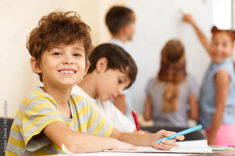 Side view of two little pupils sitting together at table and writing in copybook, one looking and smiling at camera. Smart classmates in process of learning new material in classroom. Side view of two little pupils sitting together at table and writing in copybook, one looking and smiling at camera. Smart classmates in process of learning new material in classroom.
