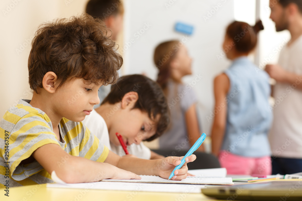 Side view of hardworking schoolboy carefully doing tasks in copybook during lesson on interesting courses. Cute little boy sitting at table and writing. Concept of modern education and learning. Side view of hardworking schoolboy carefully doing tasks in copybook during lesson on interesting courses. Cute little boy sitting at table and writing. Concept of modern education and learning.