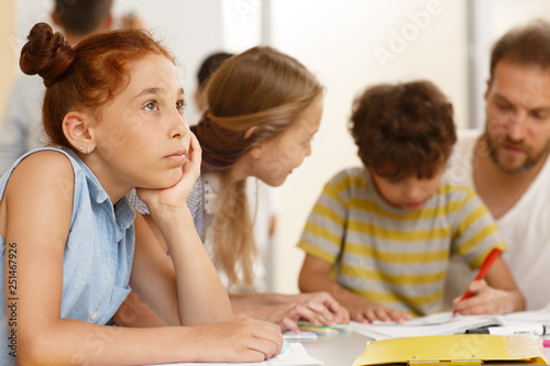 Side view of cute shoolgirl sitting at table, thinking about something and looking away in classwoom. Pupil with classmates on background learning new material with teacher on courses.