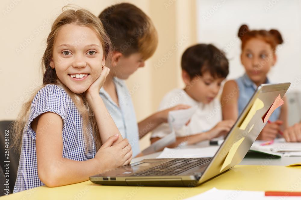 Portrait of pretty schoolgirl sitting at table, looking at camera and smiling in classroom. Cute pupil with classmates on background using laptop and network for learning. Concept of modern school.