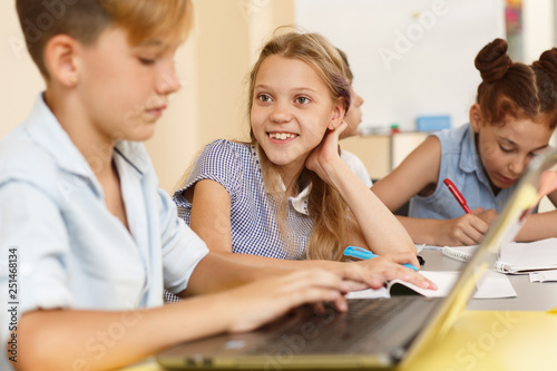 Little positive girl sitting at table, looking at boy and listening carefully. Pretty smiling schoolgirl talking with classmate using computer during lesson in school. Interesting study.