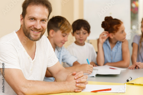 Intelligent teacher sitting at table  looking at camera and posing in school. Qualified tutor teaching little students  giving them tasks and problems in classroom. Concept of interesting education.