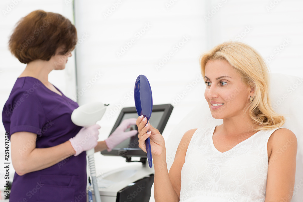 Beautiful smiling blonde sitting on coach and looking at mirror at skin after procedure in cosmetology office. Professional doctor standing with apparatus in hands on background. Concept of skin care.