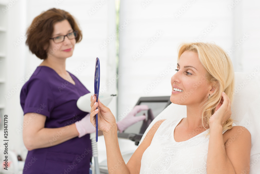 Side view of female patient satisfied with result after procedure in cosmetology office. Attractive woman looking at mirror at skin. Specialist with apparatus standing on background. Side view of female patient satisfied with result after procedure in cosmetology office. Attractive woman looking at mirror at skin. Specialist with apparatus standing on background.