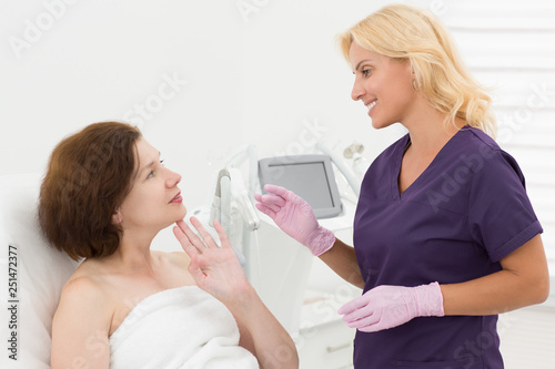 Female adult consulting with doctor cosmetologist before cosmetic procedure in cosmetology office. Woman touching with fingers face skin, profesіional beautician smiling and looking carefully.