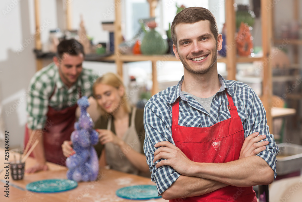 Obraz premium Portrait of handsome craftsman in checkered shirt and red apron wit folded arms looking at camera, smiling and posing during pottery class. Cheerful man enjoying new favorite hobby in art studio.