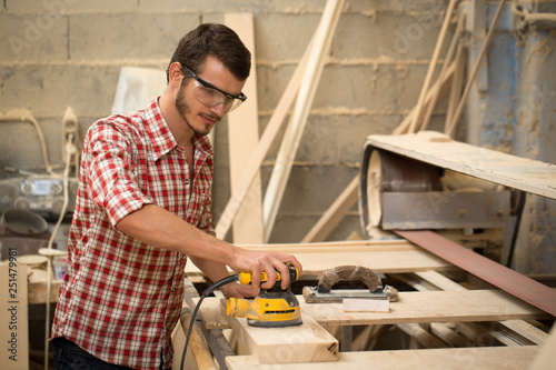 Busy and serious joiner holding yellow sander and working with wood. Professional carpenter in safety glasses standing in joiner's shop. Concept of woodworking and craftsmanship.