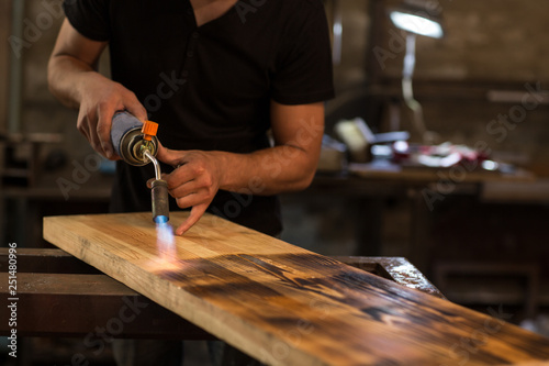Photos Crop of man wearing in black shirt, burning wooden board for displaying textures with petrol blowtorch ignition at factory