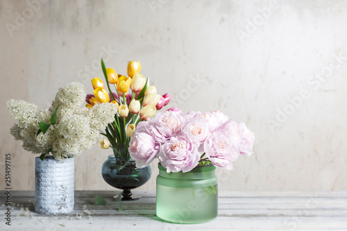 Arrangement of flowers, lilac peonies, tulips, stands on old wooden boards against one wall, in glassware of different colors