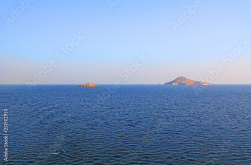 A textured background of some of the thousands of small uninhabited greek islands in the calm open water of the Agean sea with plenty of beautiful blue sky and water copy space.