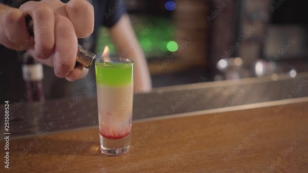 Bartender put fire from lighter to the shot with layers of differend colors of alcohol at the bar Bartender put fire from lighter to the shot with layers of differend colors of alcohol at the bar