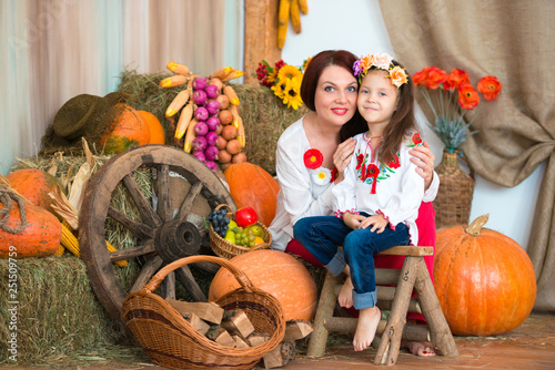 A cute, smiling mother and daughter in a colorful Ukrainian wreath and in embroidered is sitting on haystacks. Autumn decor, harvest with pumpkins