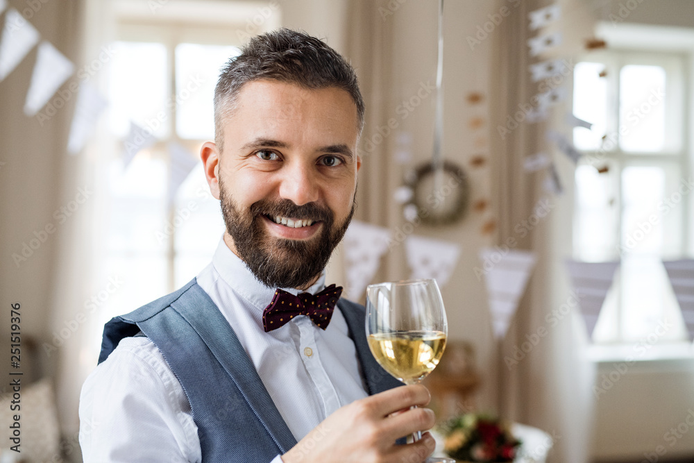 A portrait of a mature man standing indoors in a room set for a party, holding wine. A portrait of a mature man standing indoors in a room set for a party, holding wine.