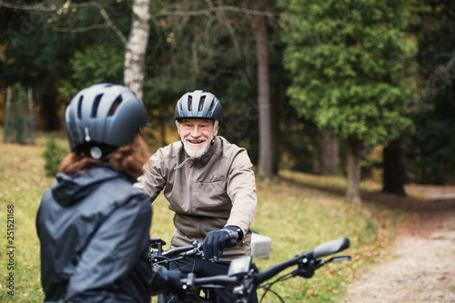 A senior couple with bicycle helmet standing outdoors on a road in park in autumn.