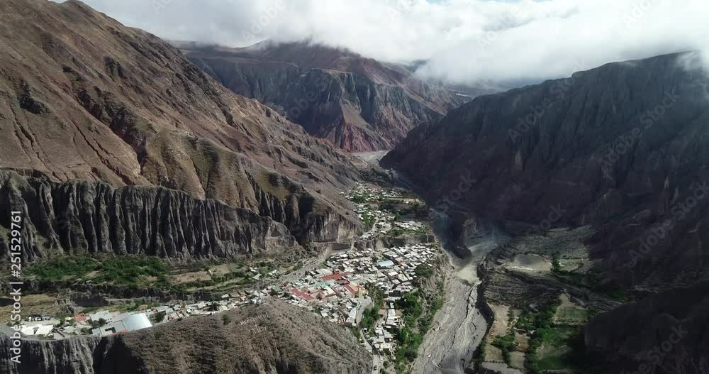 Aerial flying over Iruya, hidden town, at steep narrow valley surrounded of clouds, woods and dry colorful hills at background. Touristic town at Salta, Argentina
