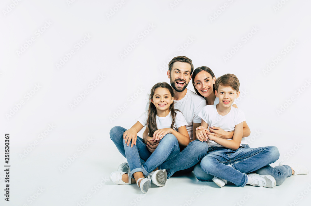 Relationship concept. Beautiful and happy smiling young family in white T-shirts are hugging and have a fun time together while sitting on the floor and looking on camera.