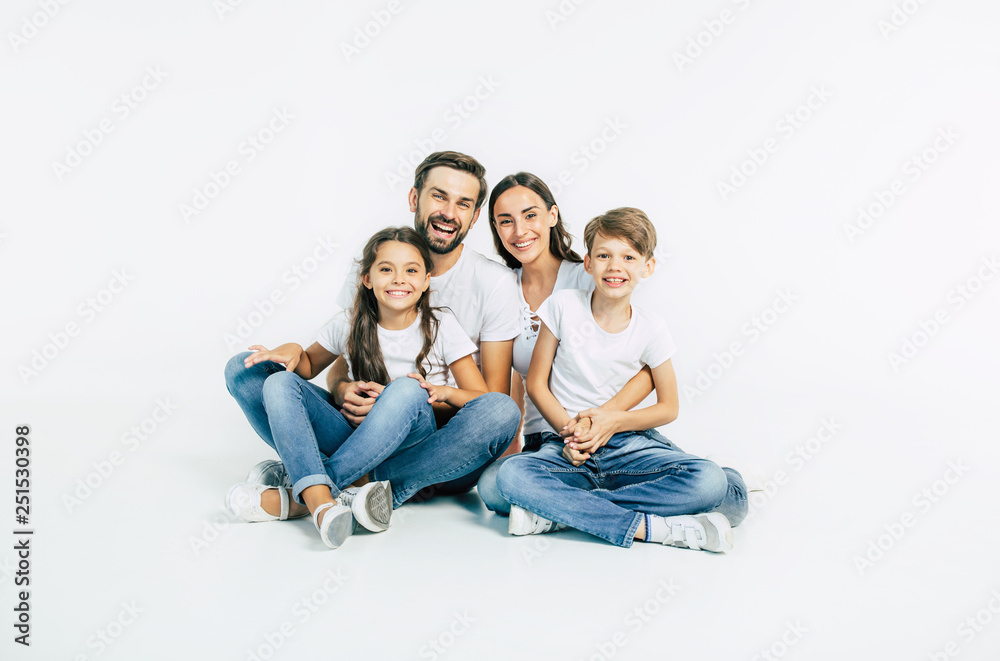 Relationship concept. Beautiful and happy smiling young family in white T-shirts are hugging and have a fun time together while sitting on the floor and looking on camera. Relationship concept. Beautiful and happy smiling young family in white T-shirts are hugging and have a fun time together while sitting on the floor and looking on camera.