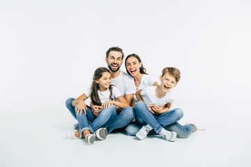 Best time with my family. Beautiful and happy smiling young family in white T-shirts are hugging and have a fun time together while sitting on the floor and looking on camera.